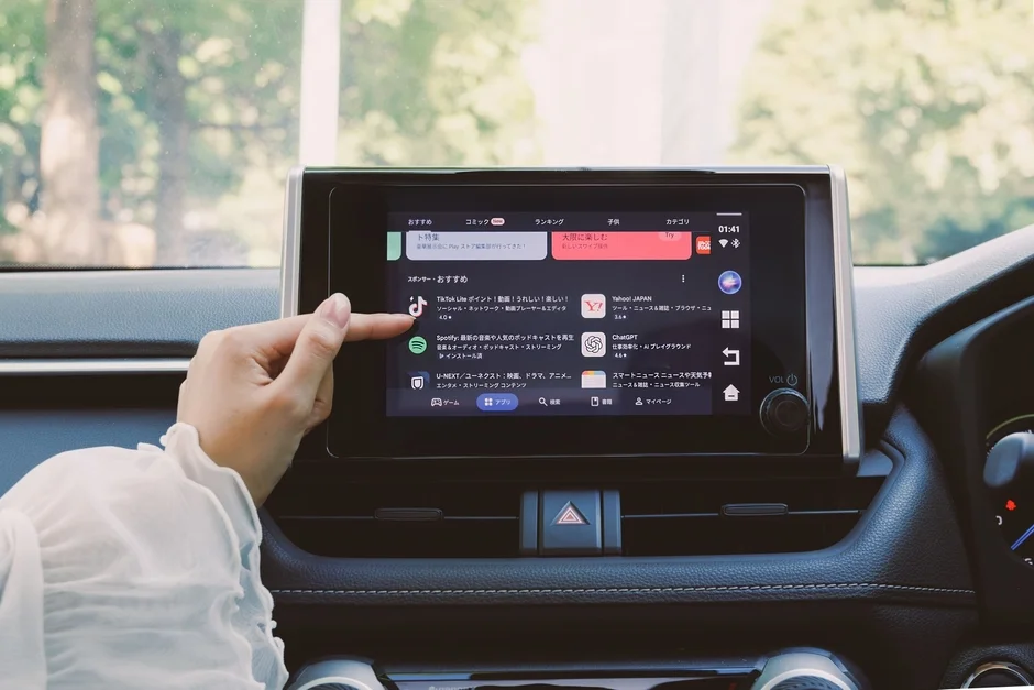 車載ディスプレイでアプリストアを操作している様子です。TikTok Lite、Spotify、ChatGPTなどのアプリが表示され、女性の指が画面をタッチしています。車内から外の緑が見える構図です。