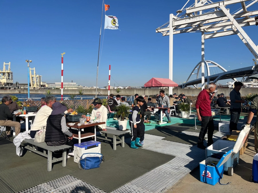 晴れた日、川沿いの屋外スペースで多くの人々がバーベキューや食事を楽しんでいます。