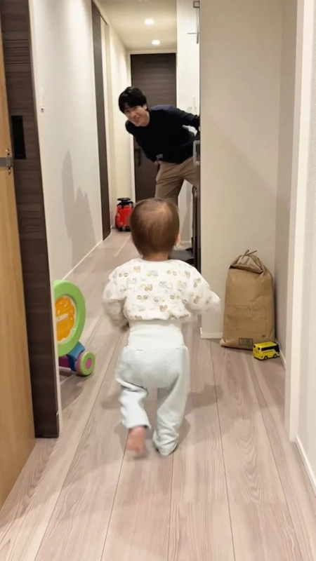 廊下で赤ちゃんが歩いており、開いたドアの奥から父親らしき男性が笑顔で覗いている