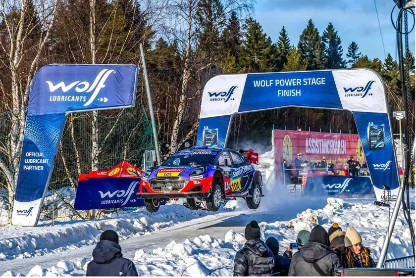 雪のラリーコースで、WOLF POWER STAGEのゴール地点を前にラリーカーが空中にジャンプする瞬間です。観客が見守る中、スポンサーのロゴが目立つアーチの下を通過する迫力あるシーンです。