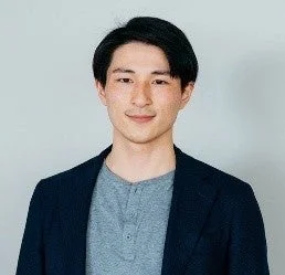 シンプルな背景の前で、ダークブルーのジャケットとグレーのTシャツを着た若いアジア人男性