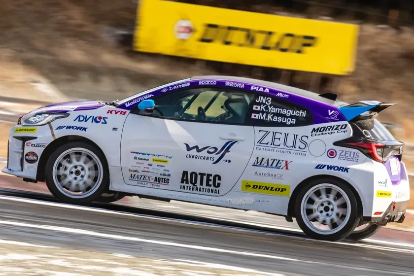 多数のスポンサーロゴが施された白いトヨタGRヤリスのラリーカーが、レーストラックを走行している。紫色のアクセントが特徴で、背景には「DUNLOP」のバナーが見える。競技中の躍動感ある瞬間を捉えた写真。