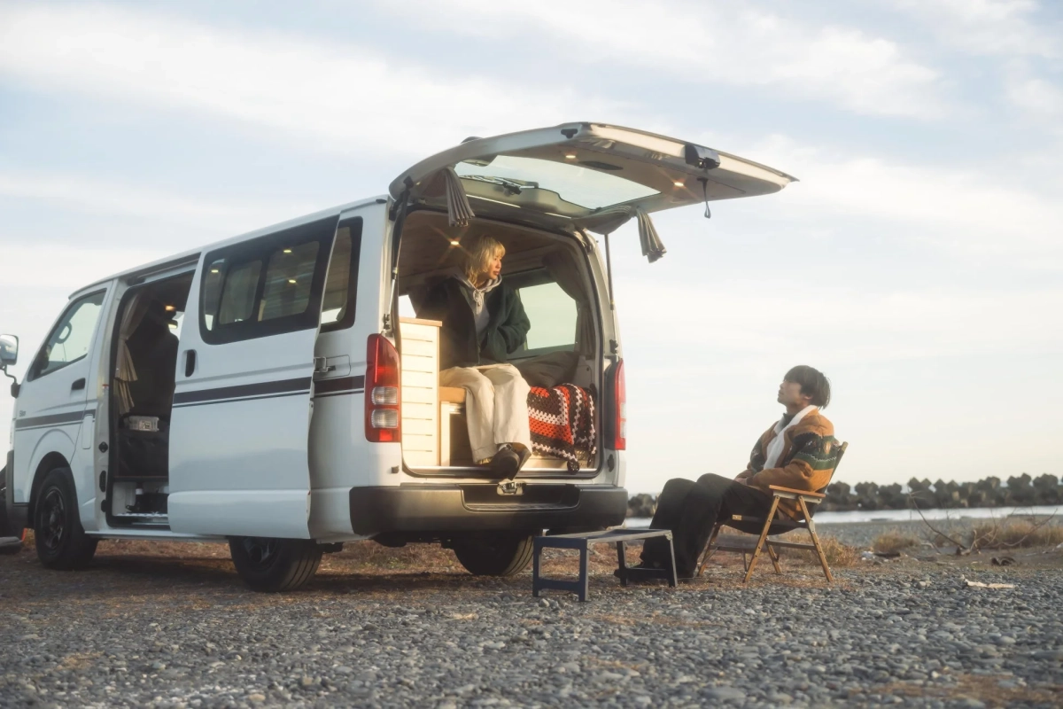 白いキャンピングカーと二人組が、砂利の広がる水辺で穏やかな時間を過ごしています。一人は車の後部座席でくつろぎ、もう一人は外で椅子に座り、景色を眺めています。