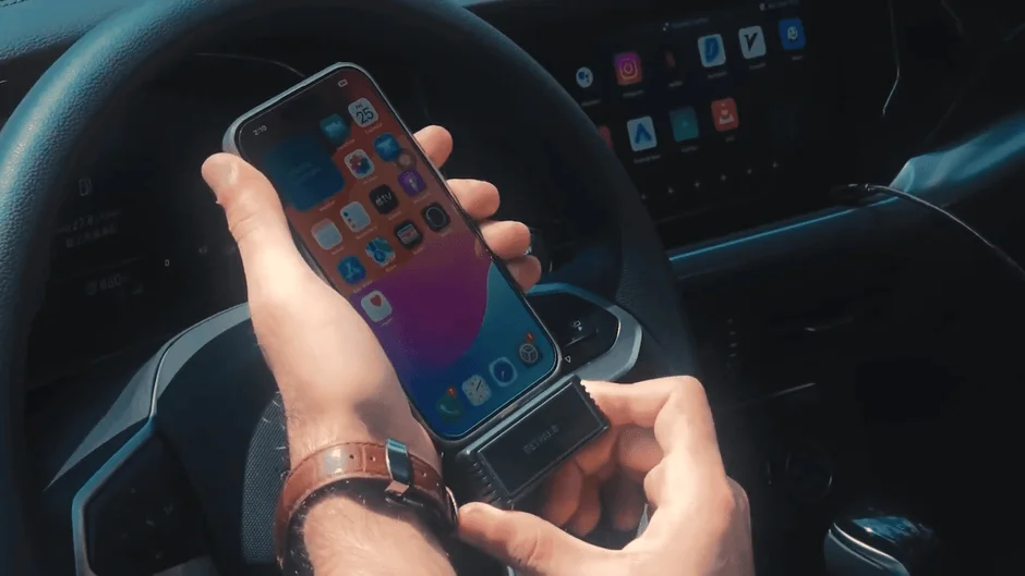 車内で、運転席に座る人物がスマートフォン（iPhone）と黒いデバイスを接続している様子。