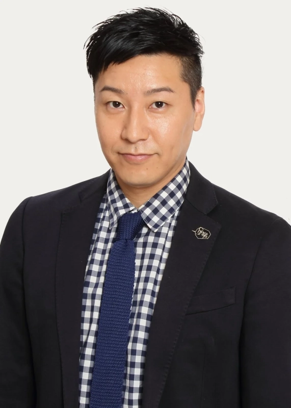 白い背景の前に立つアジア人男性の上半身ポートレート。黒いジャケットと青白のギンガムチェックシャツ、紺色のニットタイを着用し、カメラをまっすぐ見つめている。