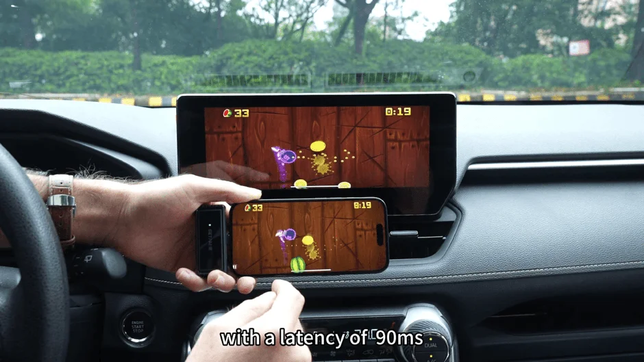 車内でスマートフォンを操作している様子が映っています。スマートフォンでプレイしているゲームがカーナビ画面にも表示されており、90msの遅延があることを示しています。
