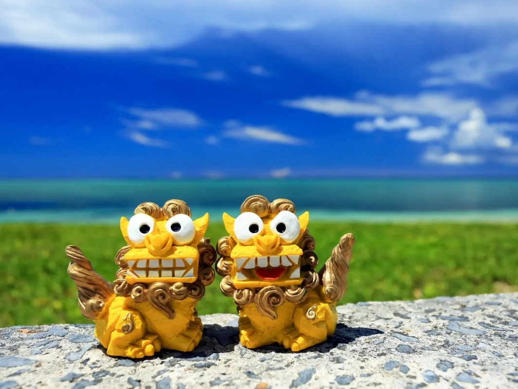沖縄の美しい青い海と空、緑の芝生を背景に、愛らしい2体の黄色いシーサーの置物が並んでいます。