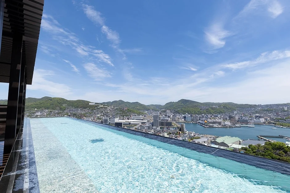 青空の下、インフィニティプールから広がる街並み、湾、そして連なる山々を一望できる絶景
