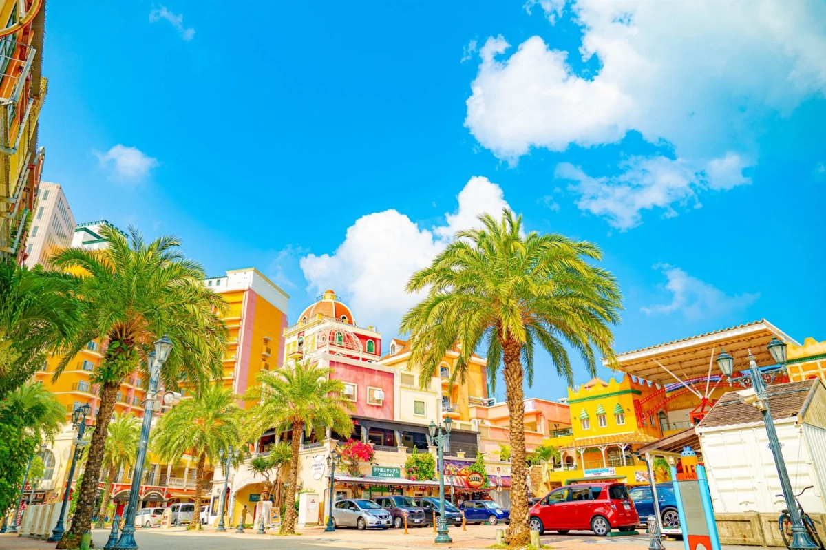 青い空と白い雲の下、ヤシの木が並ぶカラフルな建物が特徴的な街並みです。
