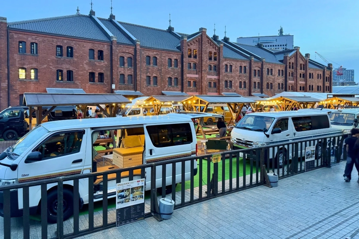 赤レンガ倉庫のような歴史的建造物を背景に、カスタムされたバンやキャンピングカーが多数展示されている屋外イベントの様子。夕暮れ時で、多くの人が会場を訪れています。