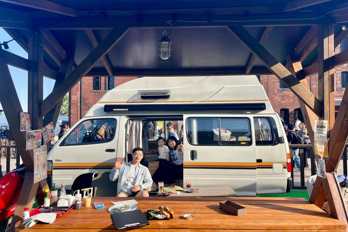 キャンピングカーの横で、笑顔の家族が屋外で食事を楽しんでいる様子。男性が手を振り、女性が子供を抱き、リラックスした雰囲気。テーブルには美味しそうな料理が並び、イベント会場のような賑わいが見えます。