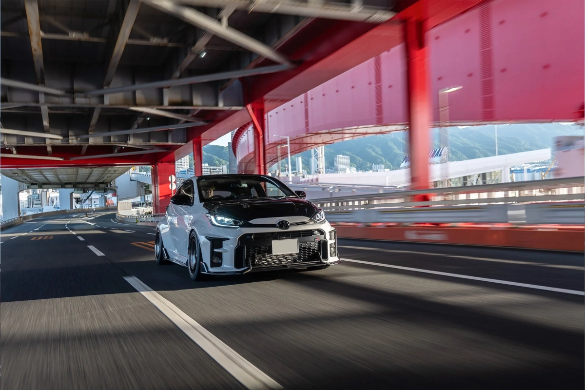 白いボディに黒いボンネットのスポーツカーが、赤い橋の下の高速道路を走行している様子。