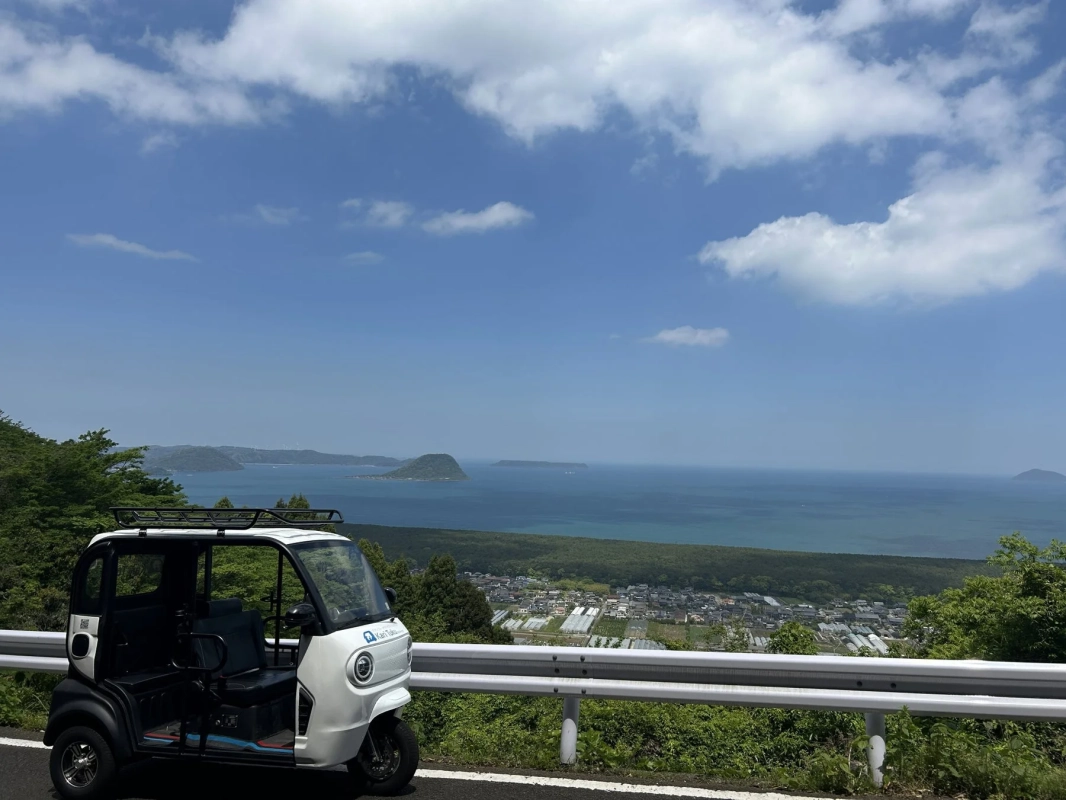青い海と島々、街並みを一望できる高台の道路に停車する白い小型電気自動車