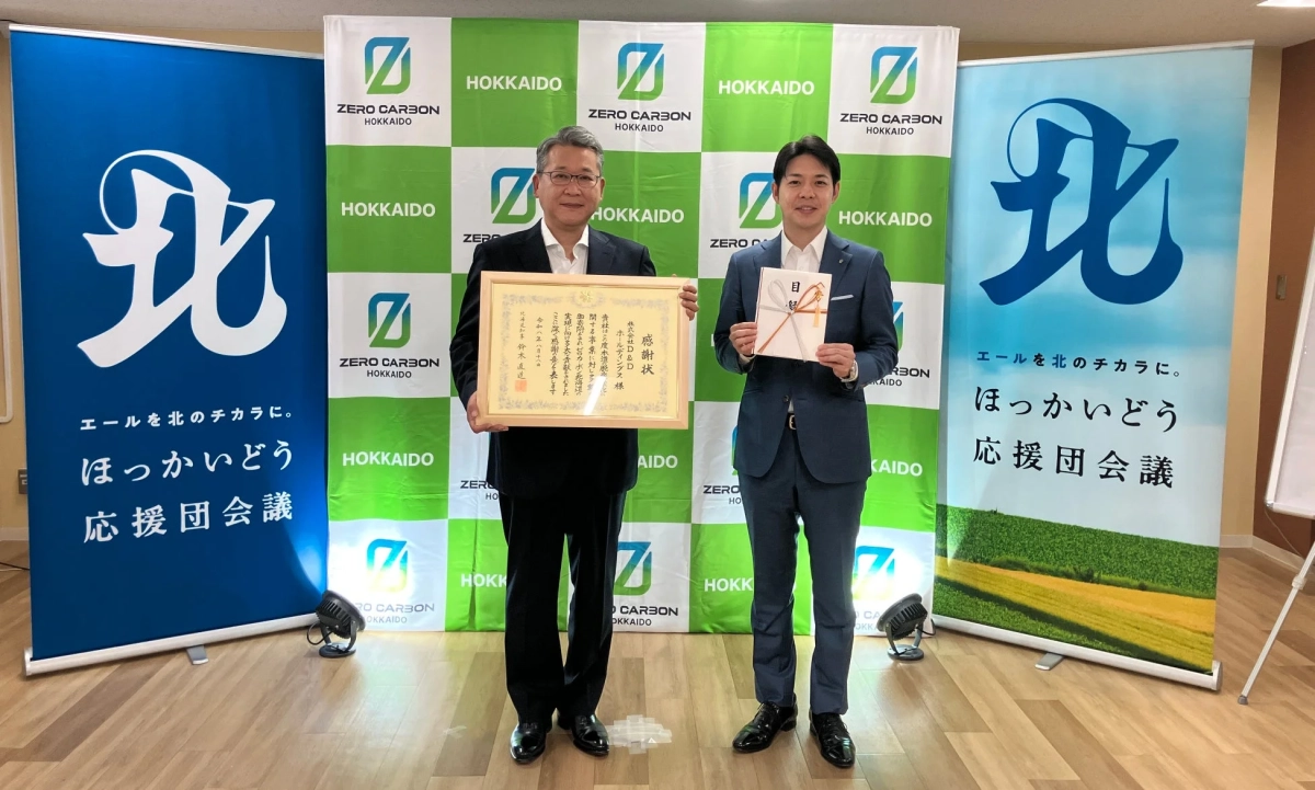 ZERO CARBON HOKKAIDOのバナーを背景に、二人の男性が感謝状と目録を持って立つ表彰式の様子。北海道知事鈴木直道の名前が見え、北海道の発展への貢献が称えられている。