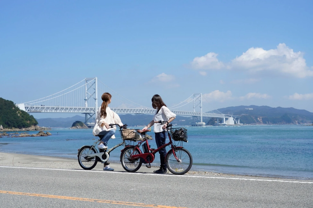 青い空と海を背景に、自転車に乗った二人の女性が橋を眺めている風景