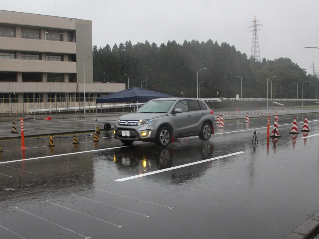 雨の中、濡れたアスファルトの路面を走行するシルバーのSUVの様子。運転訓練やテストコースでの撮影とみられ、オレンジ色のコーンや建物が背景に見えます。